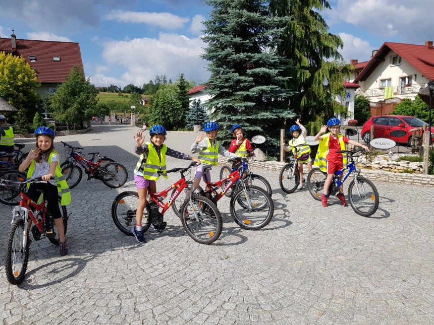 Grupa dzieci w kaskach i kamizelkach odblaskowych na wycieczce rowerowej przed pensjonatem. Dzieci siedzą na rowerach górskich i machają do aparatu na brukowanym podjeździe.