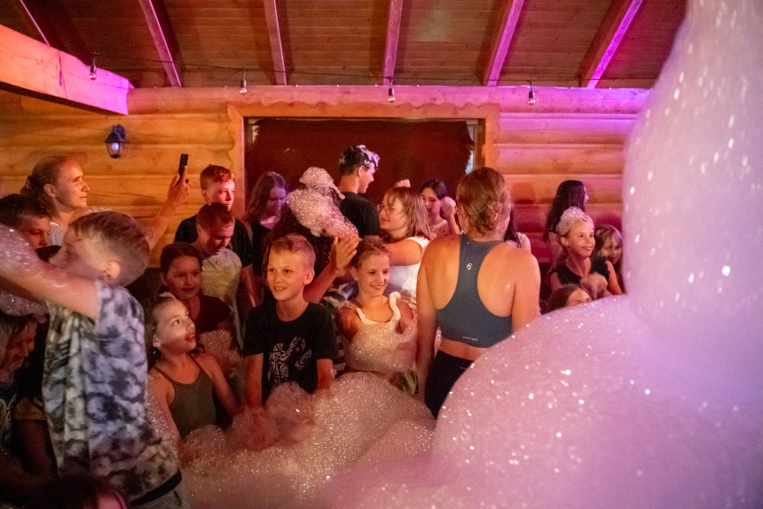 Impreza z pianą (foam party) zorganizowana dla grupy dzieci i młodzieży w drewnianym budynku. Uczestnicy bawią się w dużej ilości białej piany, rozświetlonej różowym i ciepłym światłem reflektorów.